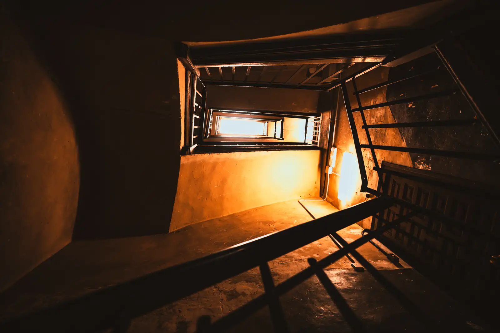 Blick von unten durch ein Treppenhaus nach oben: mehrere Stockwerke mit Geländern, warm beleuchtete Wände und ganz oben eine helle Öffnung.