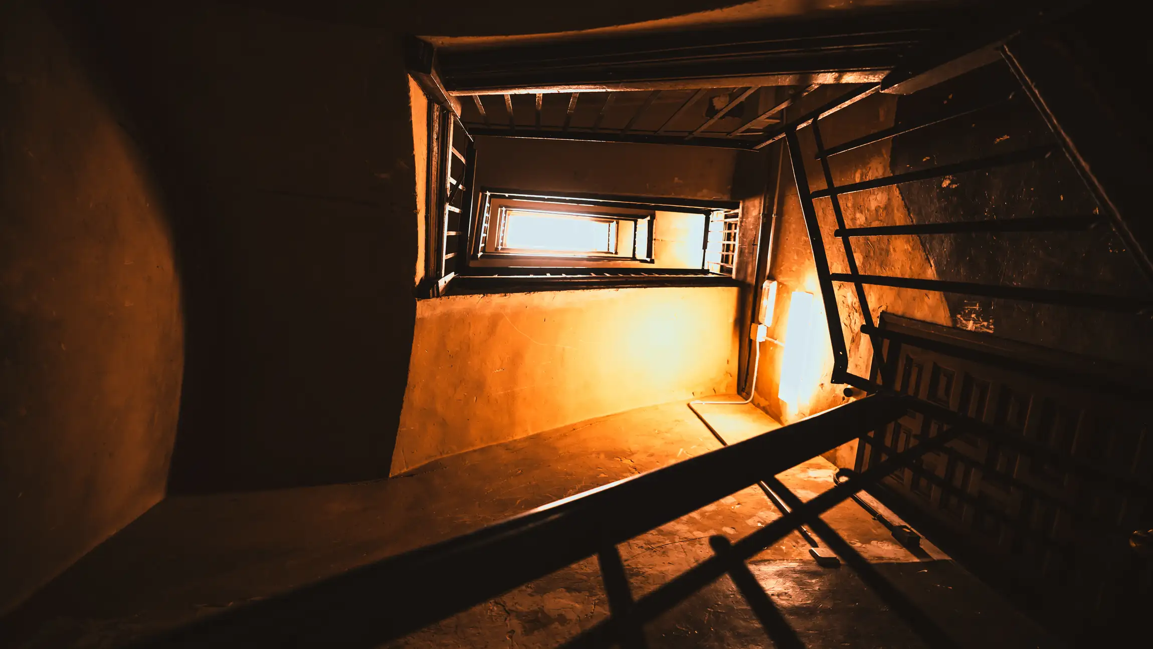 Blick von unten durch ein Treppenhaus nach oben: mehrere Stockwerke mit Geländern, warm beleuchtete Wände und ganz oben eine helle Öffnung.