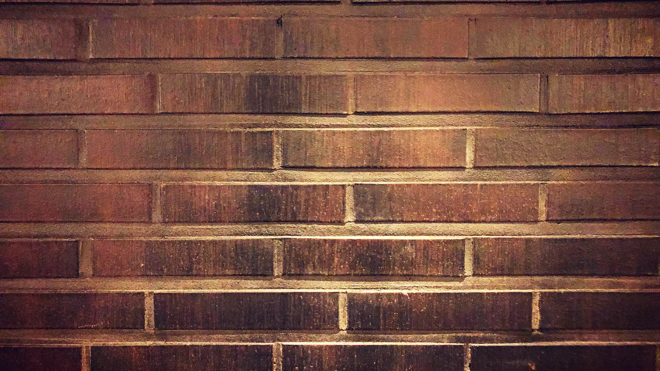 Eine Mauer aus dunkelroten Ziegeln, Reihe für Reihe versetzt gemauert, mit hellen Fugen und einem warmen Lichtstreifen quer über der Mitte.